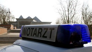 Auf dem Bild sieht man das Dach eines Einsatzfahrzeugs mit einem Blaulicht und der Aufschrift "NOTARZT". Im Hintergrund ist ein Gebäudekomplex zu erkennen, umgeben von Bäumen. Das Bild vermittelt eine Notfallsituation.