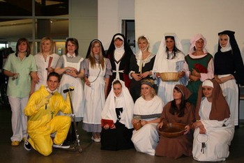 Eine Gruppe von Menschen in historischen Kostümen steht in einem Raum. Die Personen sind in verschiedenen traditionellen Kleidungsstücken gekleidet, darunter Nonnengewänder und Mittelalterkleider. Einige halten Körbe oder andere Gegenstände in den Händen. Ein Mann in einem gelben Anzug sitzt mit einem Gehstock.