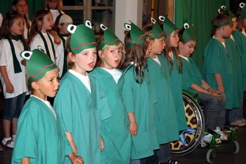 Eine Gruppe von Kindern steht in einer Reihe und trägt grüne Gewänder sowie Froschkappen. Sie wirken begeistert und singen zusammen. Im Hintergrund sind weitere Kinder und eine Person im Rollstuhl zu sehen.