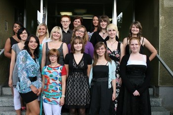 Eine Gruppe von 15 Personen steht lächelnd auf einer Treppe vor einem Gebäude. Die Frauen tragen verschiedene Outfits, von casual bis elegant. Es herrscht eine fröhliche Stimmung, und alle scheinen sich zu amüsieren.