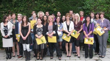 Eine Gruppe von etwa 20 Personen steht zusammen im Freien, lächelt und hält Urkunden in den Händen. Die Anwesenden sind frauen- und männergemischt, in verschiedenen Kleidungsstilen, und scheinen an einer Feier oder Zeremonie teilzunehmen. Im Hintergrund sind Pflanzen zu sehen.