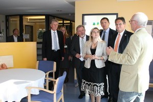 Eine Gruppe von fünf Personen steht in einem Raum. Eine Frau in der Mitte hört aufmerksam zu, während ein Mann rechts spricht. Der Raum ist hell und freundlich gestaltet, mit einem Tisch und Stühlen im Hintergrund.