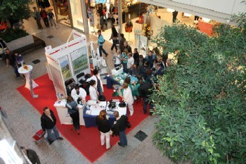 Eine Menschenmenge versammelt sich um einen Stand in einem Einkaufszentrum. Der Stand bietet Informationen und Produkte an. Roten Teppich und Pflanzen sind im Hintergrund zu sehen. Der Veranstaltungsort wirkt lebhaft und einladend.
