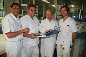 Vier Ärzte in weißen Kitteln stehen in einem Krankenhauszimmer und halten Unterlagen in den Händen. Sie wirken konzentriert und diskutieren offensichtlich ein medizinisches Thema. Im Hintergrund sind medizinische Geräte sichtbar.