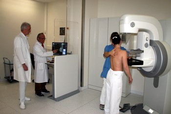 Ein Kind steht vor einem medizinischen Gerät zur Untersuchung, während ein Arzt daneben am Computer arbeitet. Eine Person in blauer Kleidung assistiert dem Kind, das sich für die Untersuchung vorbereitet. Im Hintergrund sind weitere Fachleute zu sehen.