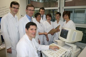 Eine Gruppe von sieben medizinischen Fachkräften in weißen Kitteln steht und sitzt um einen Ultraschallgerät. Einer der Ärzte bedient das Gerät, während die anderen auf einem Dokument schauen. Der Raum wirkt freundlich und professionell.