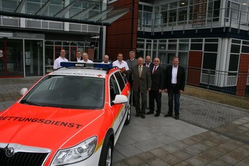 Eine Gruppe von Menschen steht vor einem Einsatzfahrzeug des Rettungsdienstes. Das Auto ist orange-weiß lackiert und trägt Licht- und Schriftzeichen. Im Hintergrund ist ein modernes Gebäude sichtbar, das in ein Programm für Rettungsdienste integriert ist.