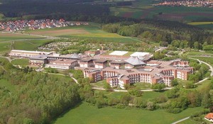 Eine Luftaufnahme eines großen, modernen Gebäudekomplexes, umgeben von grünen Bäumen und Wiesen. Im Hintergrund sind hügelige Landschaften und einige Wohngebäude sichtbar. Der Komplex hat ein auffälliges Dachdesign und mehrere angrenzende Anbauten.