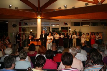 Eine Veranstaltungsbühne in einem Innenraum, auf der mehrere Personen in formeller Kleidung stehen und singen. Im Vordergrund sitzt ein Pianist. Das Publikum sitzt in Stuhlreihen und schaut aufmerksam zu. Die Atmosphäre ist festlich und gemeinschaftlich.