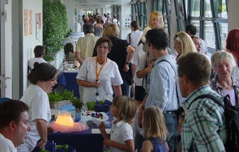 Eine lebhafte Veranstaltungsatmosphäre mit Menschen, die sich in einem Flur versammeln. Einige Personen stehen an Tischen mit Dekorationen und Pflanzen, während Kinder und Erwachsene Gespräche führen. Die Szene vermittelt ein Gefühl von Gemeinschaft und Austausch.