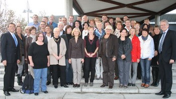 Eine Gruppe von etwa 50 Personen steht auf einer Treppe unter einem Vordach. Die Anwesenden lachen und posieren für ein Foto. Sie sind vielfältig gekleidet, einige tragen Jacken, andere Blusen oder Anzüge. Die Stimmung wirkt freundlich und gemeinschaftlich.