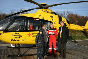 Ein roter Rettungssanitäter steht neben einem gelben Rettungshubschrauber. Er hält ein Dokument, während eine Frau und ein Junge ihm gegenüberstehen. Im Hintergrund sind Bäume und klarer Himmel zu sehen. Der Hubschrauber ist mit dem Logo des ADAC und dem Roten Kreuz gekennzeichnet.
