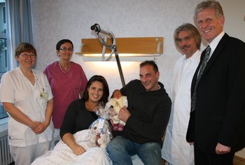 Sechs Personen stehen und sitzen in einem Krankenzimmer. Eine Frau hält ein Neugeborenes in den Armen, während der Mann neben ihr ein weiteres Baby hält. Hinter ihnen stehen zwei Krankenschwestern und zwei Ärzte. Alle lächeln und wirken zufrieden.