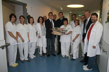 Eine Gruppe von Pflegekräften und Ärzten steht in einem Krankenhausflur. In der Mitte hält eine Person einen großen Blumenstrauß. Alle lächeln und wirken freundlich. Die Atmosphäre ist festlich und herzlich.