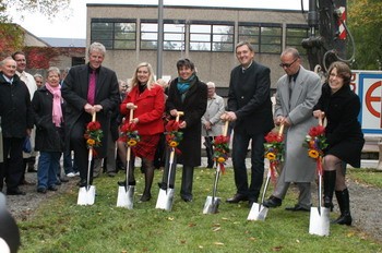 Eine Gruppe von fünf Personen steht mit Schaufeln in der Hand in einer Gartenlandschaft, bereit, einen Spatenstich zu vollziehen. Sie tragen festliche Kleidung und halten Blumensträuße. Im Hintergrund sind Zuschauer und ein Gebäude zu sehen, was auf eine öffentliche Veranstaltung hinweist.