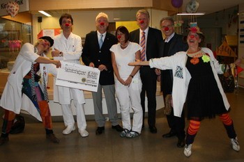 Eine Gruppe von fünf Personen steht in einem Raum, einige tragen weiße Kittel und Clownsnasen. Sie präsentieren einen großen Scheck, der auf eine Spende hinweist. Die Atmosphäre ist fröhlich, und die Clowns zeigen lebhafte Gesten, um Freude über die Spende auszudrücken.