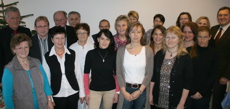 Eine Gruppe von 22 Personen steht in einem Raum und posiert für ein Gruppenfoto. Die Mehrheit trägt formelle Kleidung, einige sind lässiger gekleidet. Im Hintergrund ist ein Weihnachtsbaum sichtbar. Alle Teilnehmer lächeln und blicken in die Kamera.