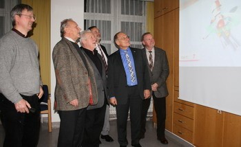 Sechs Männer stehen in einem Innenraum und schauen auf eine projizierte Präsentation. Sie scheinen interessiert und diskutieren die Inhalte, während im Hintergrund ein Bild auf der Leinwand sichtbar ist. Die Umgebung ist schlicht mit Holzregalen und Fenstern.