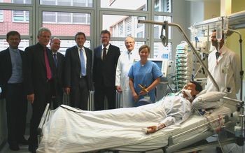 Eine Gruppe von Fachleuten und Besuchern steht zusammen in einem Krankenhauszimmer, während ein Patient im Bett liegt. Die Anwesenden zeigen Interesse und Engagement, wobei einige in medizinischer Kleidung sind. Das Bild vermittelt eine Atmosphäre der Unterstützung und des medizinischen Fortschritts.
