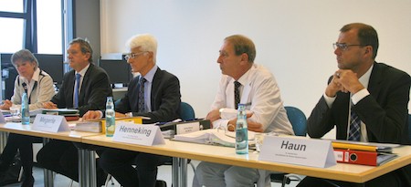 Fünf Personen sitzen an einem Tisch in einem Konferenzraum. Zwei Personen tragen Anzüge. Auf dem Tisch stehen Wasserflaschen und Ordner. Die Atmosphäre wirkt förmlich und geschäftlich, möglicherweise handelt es sich um eine Besprechung oder ein Gremium.