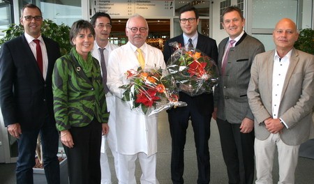 Eine Gruppe von sieben Personen steht in einem modernen Raum. In der Mitte hält ein Arzt im weißen Kittel zwei große Blumensträuße. Die anderen Personen lächeln und stehen daneben, einige in formeller Kleidung, einige in legerer Kleidung.
