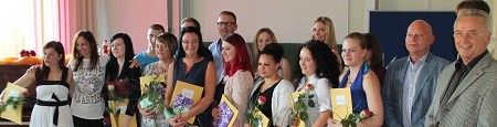Eine Gruppe von Absolventen steht auf einer Bühne und hält ihre Zertifikate in den Händen. Einige von ihnen tragen Blumen. Es gibt auch einige Personen im Hintergrund, die die Absolventen unterstützen. Die Stimmung wirkt feierlich und erfreulich.